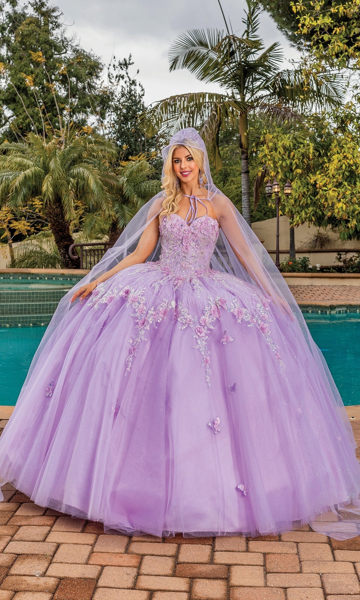 A woman stands by a pool in the Quinceanera Dress 1858 By Dancing Queen, a lilac gown with floral embellishments and a matching hooded cape, surrounded by palm trees.